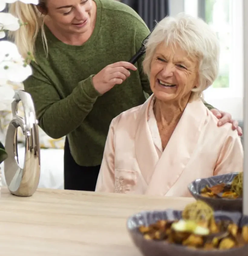 female-carer-green-top-brushing-elderly-lady-hair-infront-of-mirror-smiling-bedroom_2x3